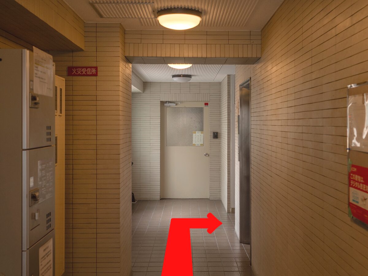 建物に入ってすぐの通路。左手に集合ポスト。奥の白いドアの手前を、赤い矢印のとおり右へ曲がる