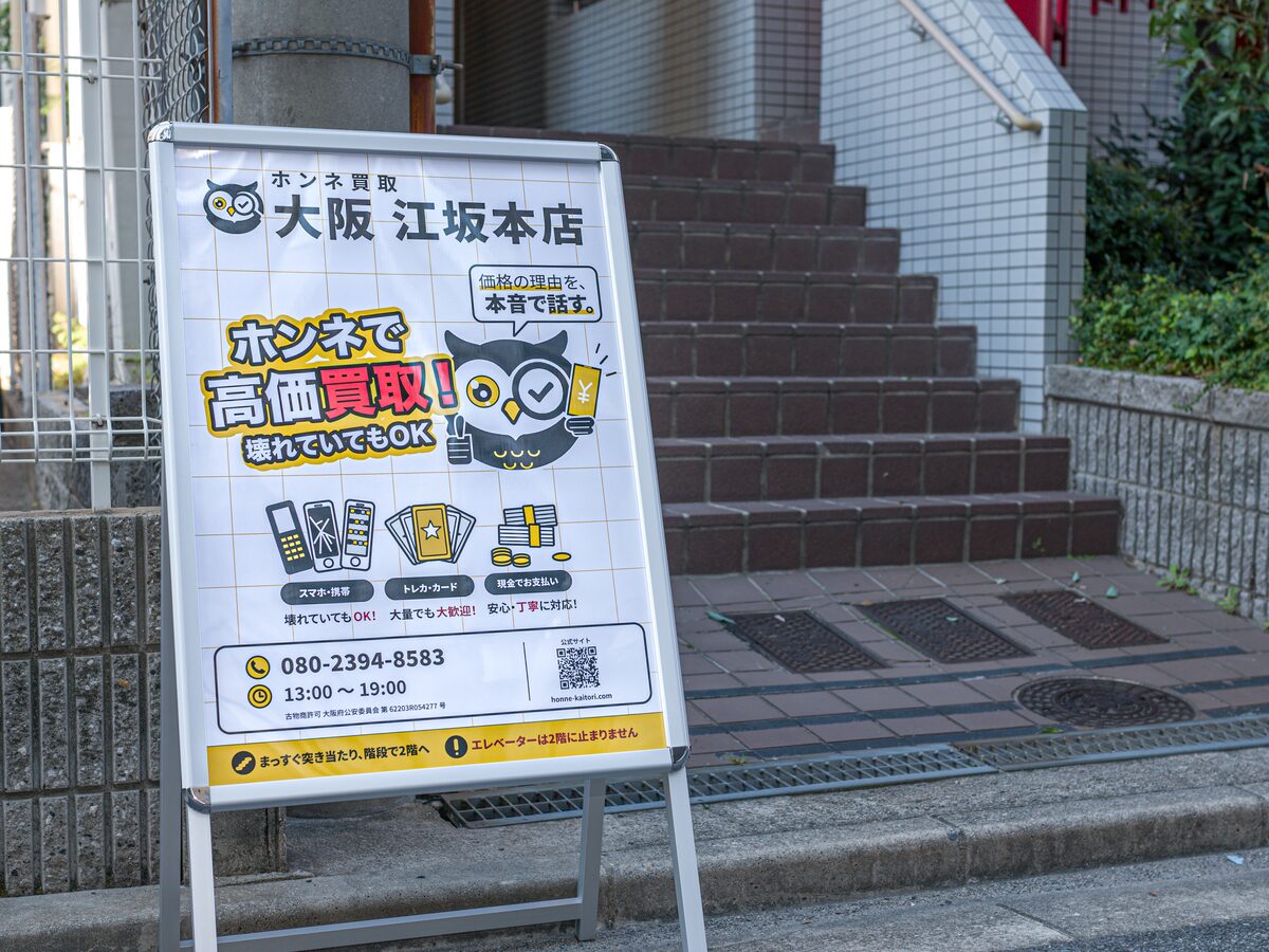 建物入口の階段の前に置いた「ホンネ買取 大阪 江坂本店」の立て看板。下の帯に「まっすぐ突き当たり、階段で2階へ」